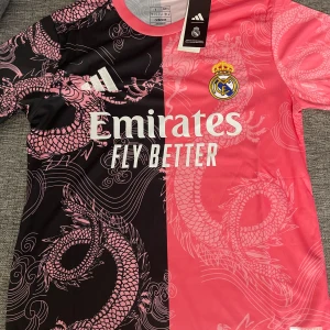 Real Madrid x Adidas rosa/svart draktröja - Säljer en unik Real Madrid fotbollströja från Adidas i rosa och svart med feta drakmönster över hela tröjan. Halva tröjan är svart med rosa drake, andra halvan är rosa med ljusare drakdetaljer. Broderad Real Madrid-logga och Adidas-märke, samt Emirates-tryck på bröstet.