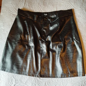 Svart kjol i skinn storlek M - Snygg svart kortkjol från Blush by Zizzi i skinnimitation med croco-mönster. Kjolen har två knappar framtill och en cool glansig finish. Storlek M