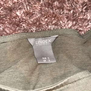 Olivgrön t-shirt med knyt från Lindex - Snygg olivgrön t-shirt från Lindex i storlek S. T-shirten har korta ärmar, rund halsringning och coola knytband i sidorna som ger en unik touch. Perfekt för dig som vill ha en enkel men trendig look.