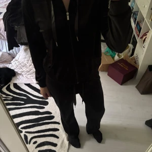 Mjukissett från Gina Tricot - Svart mjukissett i mjuk sammet från Gina Tricot. Byxorna har normal passform, raka ben och dragsko i midjan med breda satinband. Perfekta för chill dagar och riktigt sköna att ha på sig. Både zip hoodien och byxorna är i storlek M.