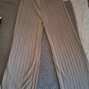 Beige plisserade wide byxor från Asu - Säljer ett par beige plisserade byxor från Asu med bred passform och elastisk midja. Byxorna har en snygg ribbad struktur och är perfekta för en chill och trendig look. Materialet känns mjukt och följsamt mot huden. Aldrig använda