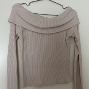 Ljusbeige ribbad offshoulder topp - Säljer en ljusbeige ribbad topp med offshoulder-design och långa ärmar. Toppen har en bred krage som vilar snyggt över axlarna och är tillverkad i mjukt, stretchigt material. Perfekt för dig som gillar stilrena och trendiga plagg med en twist.