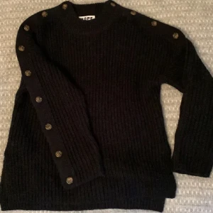 Svart stickad tröja från HOPE - Säljer en svart, chunky stickad tröja från HOPE med grov ribbstickning och snygga knappar längs axel och ärm. Tröjan är i ull- och alpackablandning, har rund hals och lång ärm. Perfekt för dig som gillar en stilren men unik look.