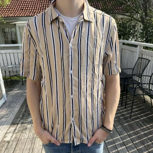 Randig skjorta - Säljer en beige skjorta med svarta och vita ränder. Skjortan har korta ärmar, klassisk krage och knäppning framtill. Perfekt för varma dagar och ger en chill vibe till din outfit.