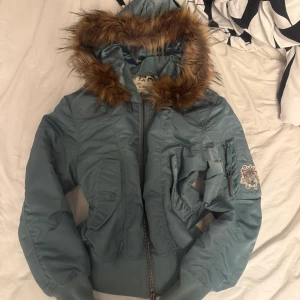 Blå bomberjacka Odd Molly med fuskpäls - Snygg blå bomberjacka från Odd Molly med stor huva och brun fuskpäls. Jackan har dragkedja, ribbade muddar och coolt broderat märke på ärmen. Fodret är mönstrat och jackan har flera fickor. Perfekt för kalla dagar och riktigt trendig look.