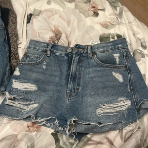Blå jeansshorts från Pull&Bear - Snygga blå jeansshorts från Pull&Bear med slitna detaljer och fransig kant. Modellen är högmidjad med klassiska fem fickor och knapp- samt dragkedja framtill. Perfekt för en chill och trendig sommarlook.