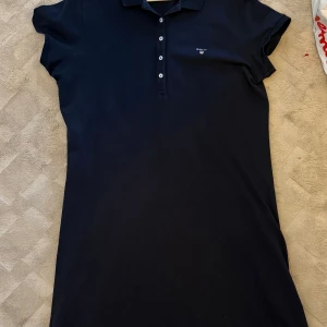 Säljer Svart pikéklänning från GANT! - Klänningen är som helt ny och endast använd ett fåtal gånger.  Storlek: XL Snygg Svart pikéklänning från GANT med klassisk krage och korta ärmar. Klänningen har knappar framtill och ett diskret broderat GANT-logo på bröstet. 