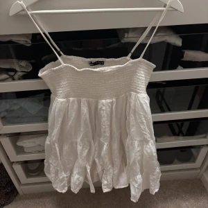 Vit somrig kortklänning med volanger - Superfin vit kortklänning från Zara med smala axelband och volangig kjol. Smockad överdel ger en skön passform och den luftiga bomullen gör den perfekt för varma dagar. Enkel och stilren design som känns riktigt fräsch.