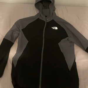 Svart och grå vindjacka från The North Face - Snygg svart och grå vindjacka från The North Face med huva och hel dragkedja framtill. Jackan har långärmade ärmar och logga på bröstet. Perfekt för dig som gillar stilren och sportig look.