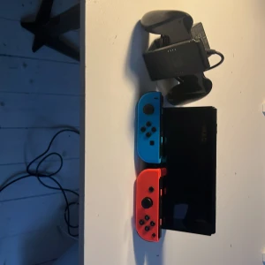 Nintendo switch  - Bilderna visar en Nintendo Switch-spelkonsol med blå och röd Joy-Con, laddare och handkontrollshållare. Perfekt för gaming på språng eller hemma, och passar dig som vill ha kul med vänner eller spela solo. En ikonisk konsol för alla som älskar spel och underhållning.