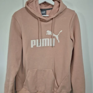 Rosa/Beige hoodie från Puma med logga - Säljer en rosa/beige hoodie från Puma med stor vit logga framtill. Tröjan har huva med snörning, magficka och ribbade muddar vid ärmslut och nederkant. Perfekt för en chill och sportig stil.