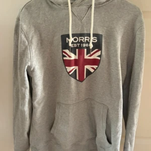 Grå hoodie från Morris med tryck - Grå hoodie från Morris med Union Jack-tryck och text på bröstet. Tröjan har huva med vita snören och en stor magficka. Tillverkad i mjuk bomull, perfekt för chill dagar. Klassisk och stilren design som funkar året runt.