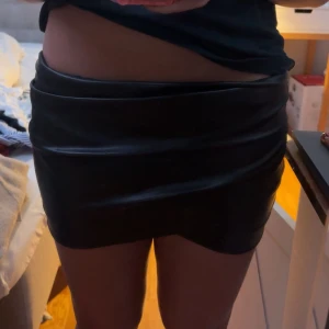 Svart skinnimitation minikjol - Säljer en svart minikjol i skinnimitation med tight passform och snygg omlott-look framtill. Kjolen har dragkedja bak och en stilren, clean design som ger en edgy vibe. Perfekt för dig som vill sticka ut med en trendig och cool look.