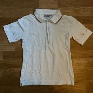 Vit piké med dragkedja - Vit pikétröja med korta ärmar och dragkedja framtill. Tröjan har broderade detaljer på ärmar och rygg samt en krage med beige kant. Liten fläck på ena armen men har inte testat att ta bort