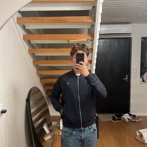 Mörkblå bomberjacka från GANT - Snygg mörkblå bomberjacka från GANT med klassisk passform. Jackan har dragkedja framtill, ribbade muddar och diskret GANT-logga broderad på bröstet. Perfekt för dig som gillar stilren och enkel design. Materialet känns lätt och smidigt.