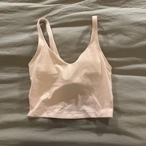 Ljusrosa linnetopp från Lululemon - Säljer en ljusrosa linnetopp från Lululemon med smala axelband och v-ringning. Toppen är croppad och har en stilren design med diskret logga på ryggen. Perfekt för träning eller chill dagar. Mjukt och stretchigt material som sitter skönt mot kroppen. Har som en inbyggd bh och har även kort som man får när man köper toppen. 
