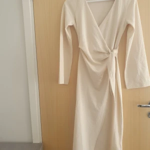 Beige omlottklänning med knyt - Säljer en lång beige omlottklänning med v-ringning och knyt i midjan. Klänningen har långa ärmar och är tillverkad i ett mjukt, lätt material som ger en snygg och enkel look. Perfekt för dig som gillar stilrena plagg med fina detaljer.