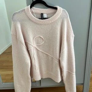 Ljusrosa stickad tröja från H&M Divided - Supermjuk ljusrosa stickad tröja från H&M Divided med rund halsringning och långärmad passform. Tröjan har en snygg ribbad kant och en dekorativ snodd i form av en rosett framtill. Perfekt för lager-på-lager under kyliga dagar.