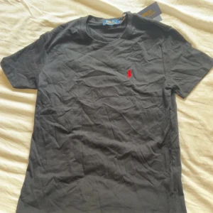 Svart t-shirt Polo Ralph Lauren S - Svart t-shirt från Polo Ralph Lauren i mjuk bomull. Klassisk passform med korta ärmar och rund hals. Liten röd broderad logga på bröstet ger en clean och stilren look. Perfekt till jeans eller shorts för en enkel men snygg outfit.