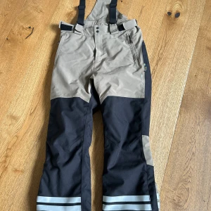 Skidbyxor Lindberg Colden 140 - Lindberg Colden skidbyxa 140 Jättefina, avtagbara hängslen, fickor på sidorna, reflexer nertill. Enda skavank är en några millimeter stor reva strax ovanför benslutet - se sista bilden. Svarta och beige. 15000 vattenpelare så väldigt vattentäta :-)