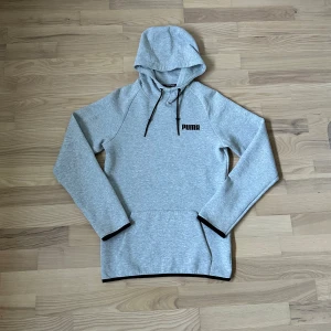 Grå hoodie från Puma med huva - Snygg ljusgrå hoodie från Puma med svart logga på bröstet. Tröjan har huva med svarta snören och en klassisk känguruficka framtill. Tillverkad i mjuk polyesterblandning, perfekt för chill eller träning. Långärmad och stilren design.