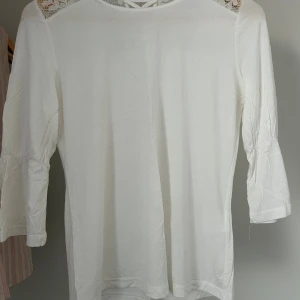 Vit blus med spets från Hellen of Sweden - Supersöt vit blus från Hellen of Sweden med trekvartsärm och fina spetsdetaljer över axlar och rygg. Blusen har ett litet snörningsparti i nacken och är gjord i mjuk bomull. Perfekt för dig som gillar romantiska och stilrena plagg.