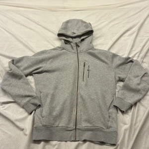 Grå hoodie från Sail Racing - Säljer en grå hoodie från Sail Racing med hel dragkedja och broderad logga på bröstet. Tröjan har huva med snörning, ribbade muddar och en praktisk bröstficka med dragkedja. 
