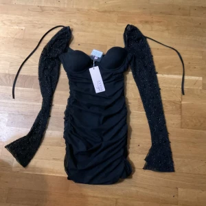 Svart glittrig kortklänning från oh polly  - Supersnygg svart kortklänning från oh polly  med figurnära passform, vadderad byst och långa glittriga ärmar med snörning. Klänningen har rynkade detaljer längs sidorna och mesh på ärmarna som ger en extra cool look. Aldrig använd helt ny men va för liten för mig nypris är 1200 säljer den för 550, finns att hämta i Karlstad Kasernhöjden.