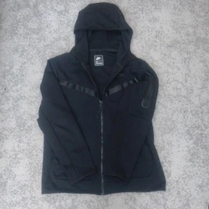 Svart Nike vindjacka med huva - Svart Nike tech i storlek M. Jackan har hel dragkedja, huva och diskreta svarta detaljer över bröstet. Praktiska fickor och en extra ficka med dragkedja på ärmen. Perfekt för dig som gillar sportig och clean stil.