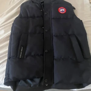 Svart dunväst från Canada Goose - Snygg svart dunväst från Canada Goose med klassisk logga på bröstet och praktiska fickor framtill. Västen har knappar och dragkedja samt quiltad design som håller värmen. Perfekt för lager-på-lager-stil under kyliga dagar.