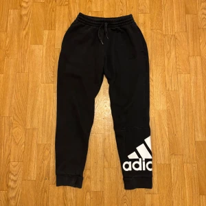 Svarta mjukisbyxor från Adidas - Svarta mjukisbyxor från Adidas med stor vit logga på ena benet. Byxorna har resår i midjan med snörning och muddar vid bensluten. Perfekta för chill eller träning, tillverkade i mjukt material för maximal komfort.