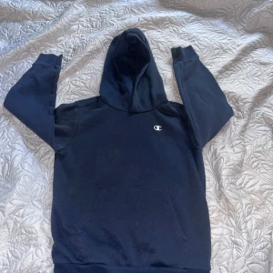 Svart hoodie från Champion - Svart hoodie från Champion med klassisk logga på bröstet. Tröjan har huva, magficka och långa ärmar. Perfekt för chill dagar eller när du vill ha en enkel och snygg look. Materialet är mjukt och skönt, troligen bomull eller polyester. Passar till 10-12 år  nypris 275
