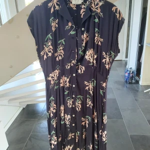 Mörkblå blommig långklänning från Emmamalena - Säljer en mörkblå långklänning från Emmamalena med kaprefolmönster i ljusrosa och grönt. Klänningen har korta ärmar, knappar hela vägen framtill och en avslappnad passform. Perfekt för dig som gillar färg och mönster!