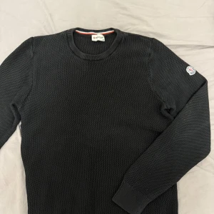 Svart stickad tröja från Moncler - Säljer en svart stickad tröja från Moncler med rund halsringning och Moncler-logga på ärmen. Tröjan har en grovstickad struktur och långa ärmar, perfekt för kyligare dagar. Klassisk och stilren design som funkar till det mesta.