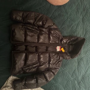 Svart Parajumpers Buzz Pufferjacka - Säljer en svart pufferjacka från Parajumpers, modell Buzz Puffers. Jackan har en glansig finish, huva och markerad quiltad design. Stängs med dragkedja och har en cool orange detalj vid nacken. Perfekt för kalla dagar och riktigt snygg till streetwear.