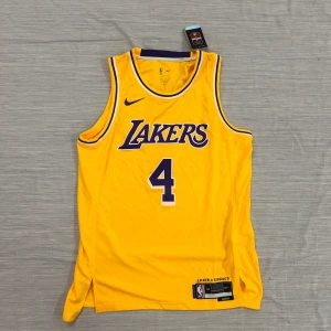 Gul Lakers baskettröja Nike L - Snygg gul Lakers baskettröja från Nike, Icon Edition med nummer 4 på bröstet. Tröjan är ärmlös och har lila och vita detaljer runt hals och ärmhål. Tillverkad i Dri-Fit meshmaterial som andas, perfekt för träning eller match. NBA Authentics-märke och texten 'Leave a Legacy' nedtill.