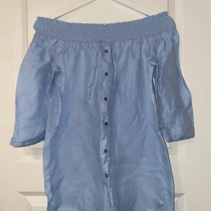 Ljusblå offshoulder klänning River Island - Säljer en ljusblå kortklänning från River Island med offshoulder-design och knappar framtill. Klänningen har trekvartsärm och är gjord i ett mjukt bomullsmaterial. Perfekt för sommarens alla tillfällen, snygg och bekväm med smock upptill.