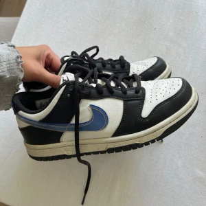 Nike Dunk Low svart/vit/blå - Nike Dunk Low sneakers i svart och vitt med blå swoosh. Klassisk siluett med svart snörning, perforerad tå och robust gummisula. Ovandel i skinn och syntet, perfekt för dig som gillar streetstyle och vill sticka ut med en ikonisk modell.