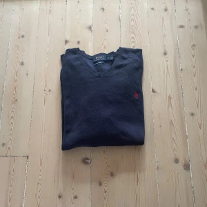 Mörkblå v-ringad tröja Polo Ralph Lauren - Mörkblå v-ringad tröja från Polo Ralph Lauren i mjuk pima bomull. Klassisk design med röd broderad logga på bröstet och ribbade muddar. Perfekt för dig som gillar stilrena och tidlösa plagg.