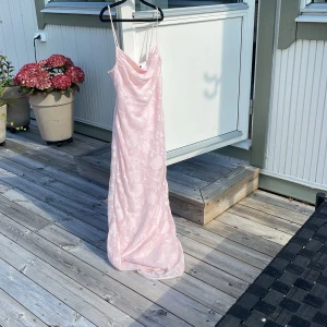 Ljusrosa långklänning från Gina Tricot - Supersöt ljusrosa långklänning från Gina Tricot med tunna axelband och draperad urringning. Klänningen har ett subtilt blommigt mönster i tyget och är tillverkad i ett glansigt, satinliknande material som ger en lyxig känsla. Har aldrig används