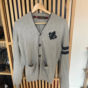 Grå Cardigan från Morris med emblem - Säljer en grå cardigan från Morris med emblem på bröstet. Den är i väldigt fint skick och är perfekt för hösten. Storlek small. 