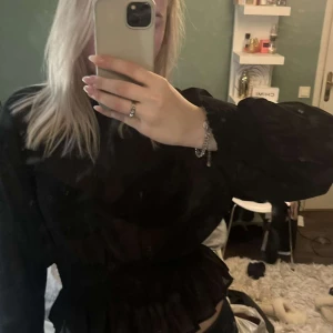 Svart volangblus med markerad midja - Säljer en svart blus i tunn, luftig polyester med långa ärmar och smockad, markerad midja. Blusen har volangdetalj nedtill och vid ärmslut, vilket ger en snygg och trendig siluett. Perfekt att styla med jeans eller kjol för en cool look.
