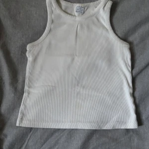 Vit ribbad linnetopp från Lindex - Säljer en enkel och stilren vit ribbad linnetopp från Lindex. Toppen är ärmlös med klassisk rund halsringning och har en tight passform. Perfekt att matcha med jeans eller shorts för en clean look.