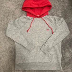 Grå och röd hoodie från Levi's XS - Snygg grå hoodie från Levi's i storlek XS med röd huva och matchande snören. Tröjan har klassisk känguruficka framtill och broderad Levi's-logga på bröstet. Perfekt för chill dagar och streetstyle. Tillverkad i mjuk bomullsmix för extra komfort. ‼️ Har hål men syns inte ‼️