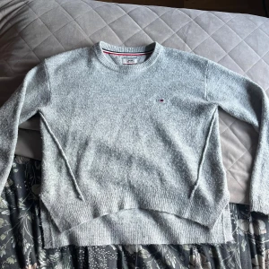Ljusgrå stickad tröja Tommy Jeans S - Mysig ljusgrå stickad tröja från Tommy Jeans med rund halsringning och diskret logga på bröstet. Tröjan har ribbade muddar vid ärmslut och nederkant, samt en klassisk passform som funkar till allt. Perfekt för kyliga dagar.