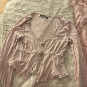 Ljusrosa omlottkofta från Gina Tricot - Superfin ljusrosa omlottkofta från Gina Tricot i mjuk bomull. Koftan har spetsdetaljer längs v-ringningen och en liten rosett framtill. Croppad passform och långa ärmar ger en söt och feminin vibe. Perfekt att styla med jeans eller kjol.