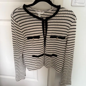 Randig kofta från H&M med gulddetaljer - Snygg randig kofta från H&M i svart och vit stickad kvalitet. Koftan har långärmad design, rund hals och dekorativa fickor med guldfärgade knappar framtill. 