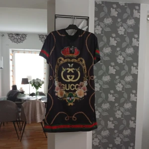 Svart kortklänning med tryck - Svart kortklänningi med färgstarkt tryck av kedjor, blommor, fjärilar och krona. Stora guldiga Gi-loggor framtill och baktill, samt röda och gröna detaljer på ärmar och nederkant. Klänningen har rund hals och korta ärmar.