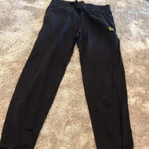 Svarta mjukisbyxor EA7 Emporio Armani - Svarta mjukisbyxor från EA7 Emporio Armani med gul logga på vänster lår. Byxorna har resår i midjan med snörning och är tillverkade i mjukt bomullsmaterial. Perfekta för en chill och sportig look.