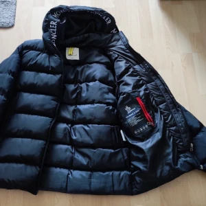 Svart Moncler dunjacka med huva - Snygg svart dunjacka från Moncler med quiltad design och huva med logotyp. Jackan har dragkedja, innerficka med röd detalj och klassisk Moncler-patch på bröstet. Perfekt för kalla dagar när du vill hålla stilen och värmen.